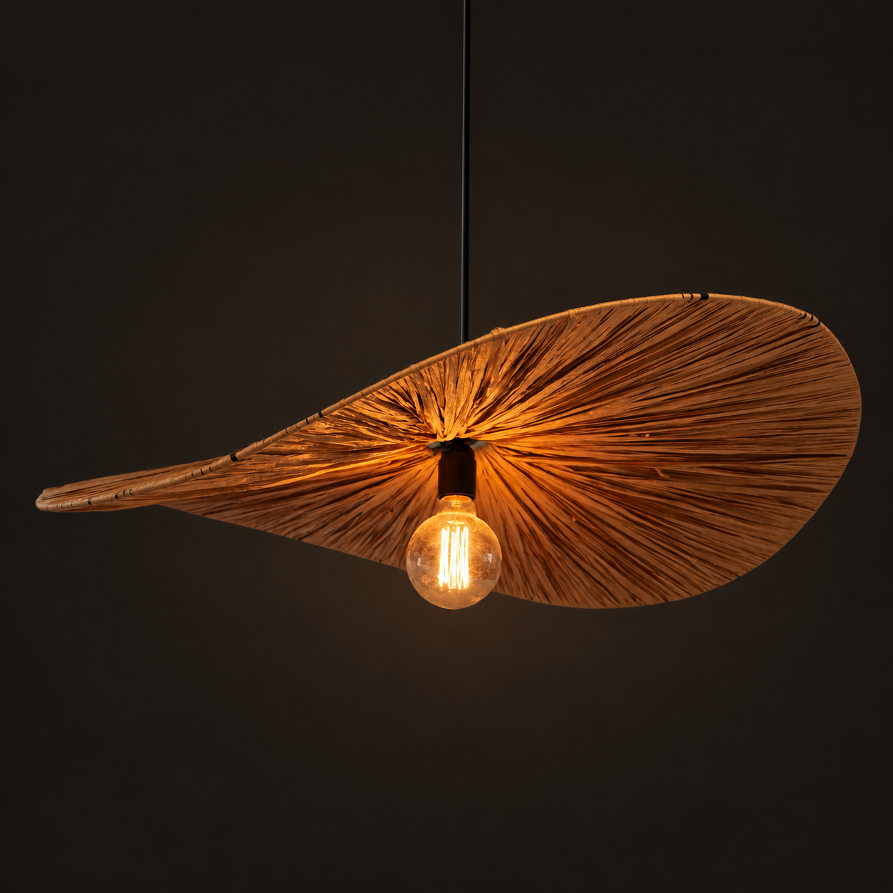 Standard Raffia Pendant Lamp illuminated, with a visible bulb beneath its folded shade against a dark background.