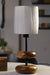 A serene forest retreat bedside features the 'Earthy Wood Glow' table lamp‚Äîteak wood base, sleek metal stem, and off-white fabric shade‚Äîon a rustic wooden table, with a golden glow illuminating books, a ceramic mug, and a lush forest view through a window