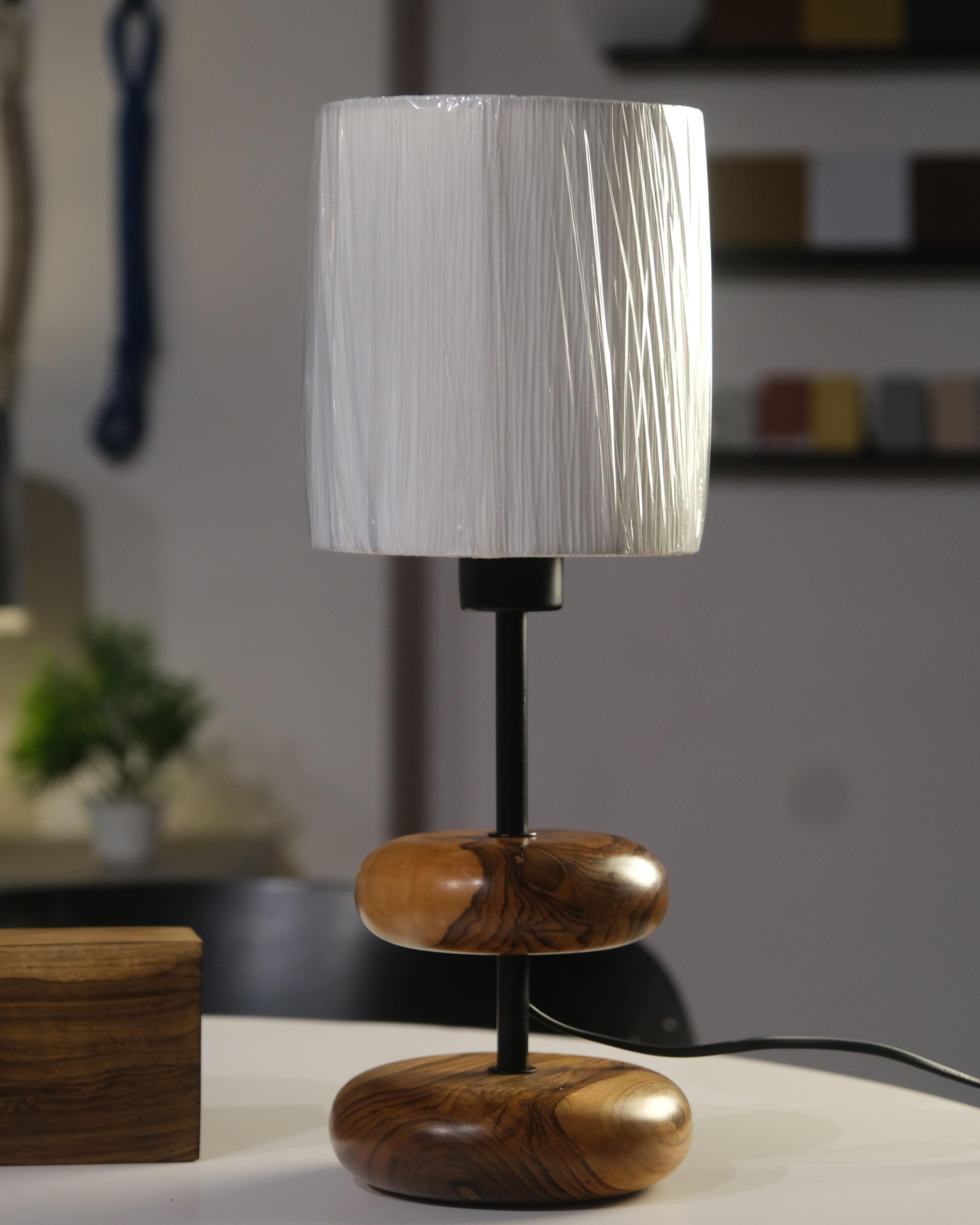A serene forest retreat bedside features the 'Earthy Wood Glow' table lamp‚Äîteak wood base, sleek metal stem, and off-white fabric shade‚Äîon a rustic wooden table, with a golden glow illuminating books, a ceramic mug, and a lush forest view through a window