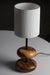 A warm teak wood table lamp with a sleek metal stem and soft off-white fabric shade glows gently on a rustic wooden bedside table in a forest retreat bedroom, surrounded by nature-inspired decor and a view of pine trees through a window.