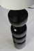Contemporary table lamp featuring a glossy black geometric base and a white cylindrical lampshade on a desk.