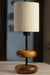 On a honey-toned wooden bedside table in a forest retreat, the 'Earthy Wood Glow' lamp with its teak wood base, metal stem, and off-white shade casts a soft light, framed by a moss-green wall, nature sketches, and a view of tall pine trees outside.
