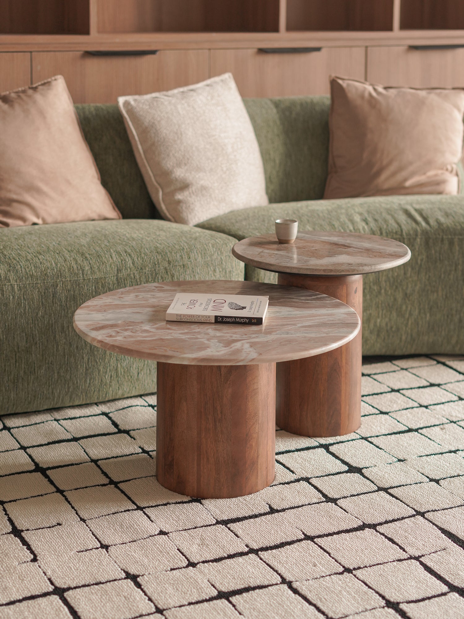 Two wooden coffee nesting tables in front of a green sofa with beige cushions on a tiled floor.