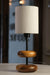 The 'Earthy Wood Glow' lamp, featuring a rich teak wood base, neutral metal stem, and off-white fabric shade, sits on a reclaimed wood bedside table in a cozy forest retreat, with books and lavender nearby, illuminated against a woodland backdrop