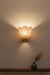 Wall-mounted lamp with a checkered pattern casting light on a plain wall, with a vase and dried plants on a shelf below.