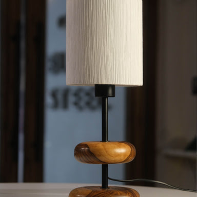 The 'Earthy Wood Glow' lamp, featuring a rich teak wood base, neutral metal stem, and off-white fabric shade, sits on a reclaimed wood bedside table in a cozy forest retreat, with books and lavender nearby, illuminated against a woodland backdrop