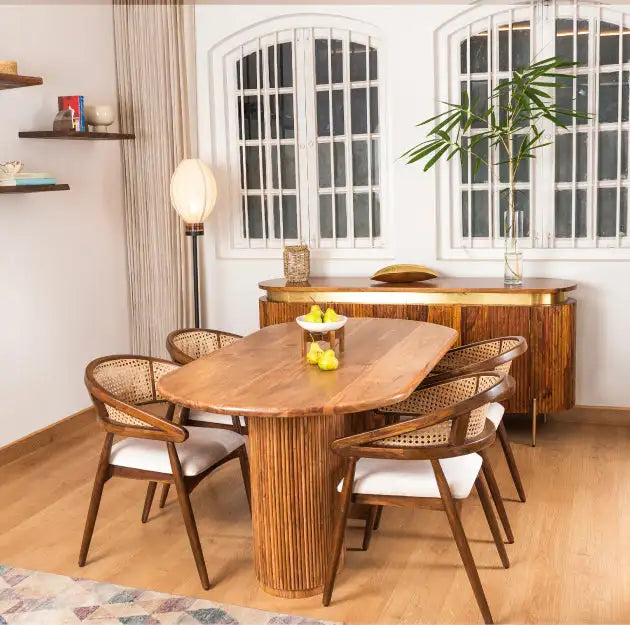 The dining table is a rustic oval wooden piece with a central pedestal base made of vertical bamboo slats, featuring a warm natural wood finish and matching woven cane chairs.
