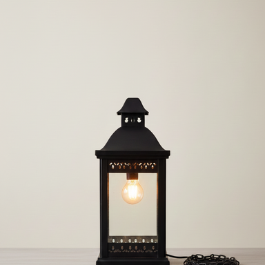Black lantern-style table lamp with a lit bulb on a white surface and light gray background