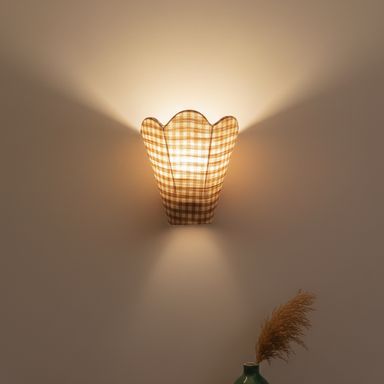 Wall-mounted lamp with a checkered pattern casting light on a plain wall, with a vase and dried plants on a shelf below.